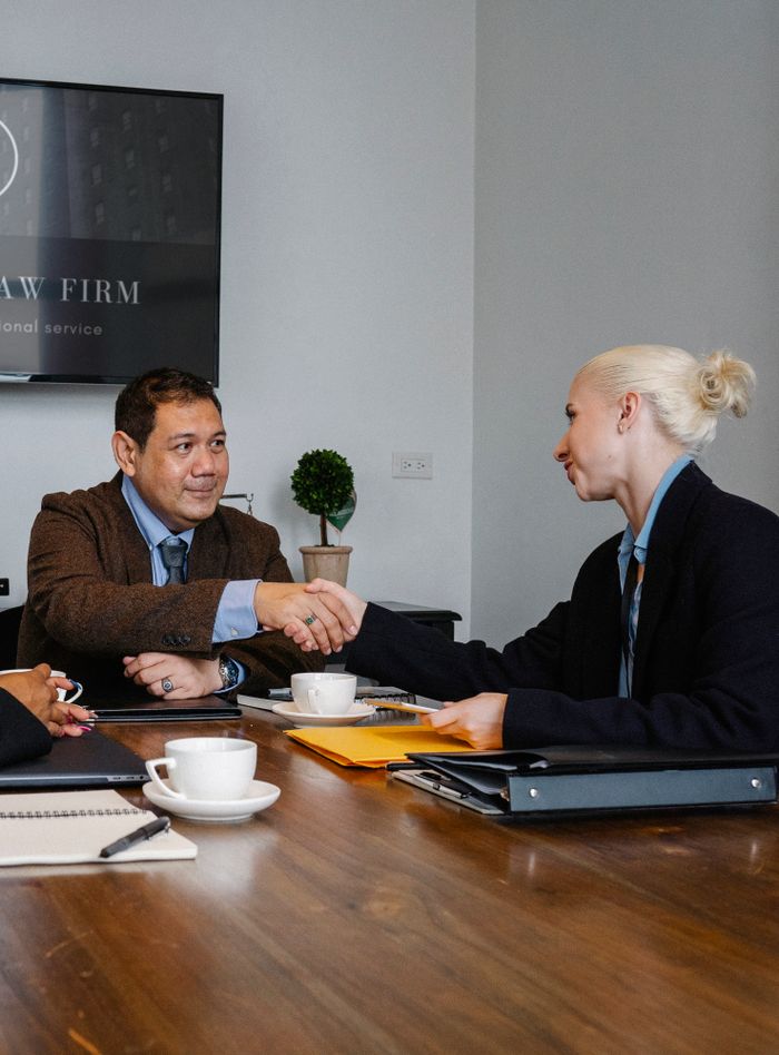 Smiling multiracial coworkers in formal clothes gathering at wooden table with cups of coffee and various folders and shaking hands while looking at each other
