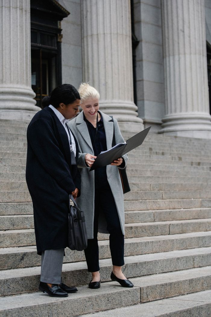 Full length of positive young multiracial female lawyers in formal outfits reading documents while standing on stairs of aged columned courthouse